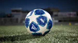 Baltas ir mėlynas futbolo kamuolys padėtas ant žalios stadiono žolės prieš svarbias rungtynes.