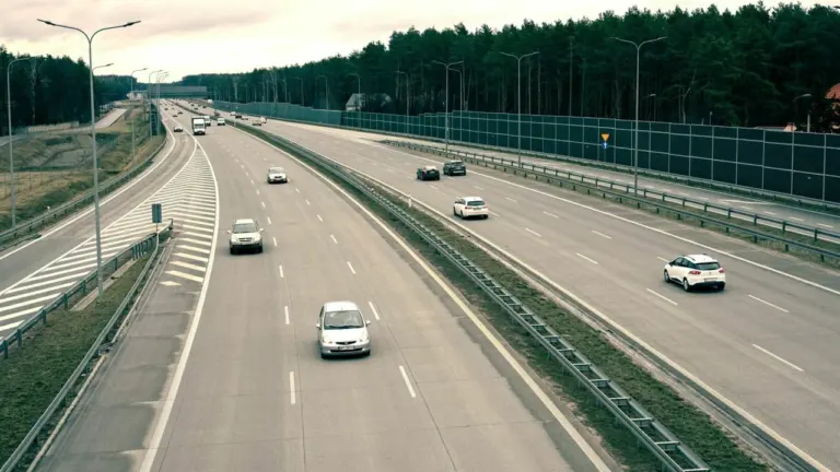Keli automobiliai važiuoja plačiu, moderniu greitkeliu šalia miško bei apsauginių garso barjerų.