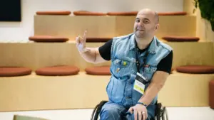 Besišypsantis vyras neįgaliojo vežimėlyje vilki džinsinę liemenę ir rodo laisvės ar pergalės ženklą.
