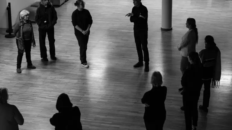 Grupė žmonių muziejaus salėje klausosi gido, kuris stovi centre ir rodo ranka.