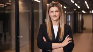 Šviesiaplaukė verslo moteris juodu švarku stovi biuro koridoriuje sukryžiavusi rankas ir žvelgia į kamerą.