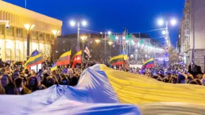 Didelė minia žmonių laiko Ukrainos vėliavą gatvėje su Lietuvos vėliavomis vakaro metu.