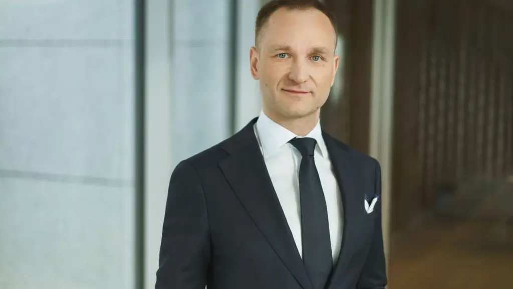Trumpai kirptas vyras šviesiais plaukais dėvi tamsų kostiumą, baltus marškinius ir tamsų kaklaraištį.