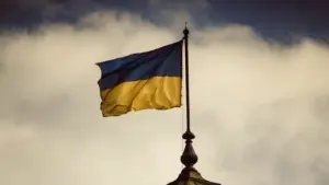 Ukrainos vėliava plazda virš pastato stogo niūriame danguje, simbolizuojanti laisvę ir pasipriešinimą.