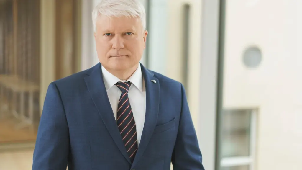 Vidutinio amžiaus žilaplaukis vyras vilkintis tamsiai mėlyną kostiumą, baltus marškinius ir dryžuotą kaklaraištį.
