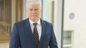 Vidutinio amžiaus žilaplaukis vyras vilkintis tamsiai mėlyną kostiumą, baltus marškinius ir dryžuotą kaklaraištį.