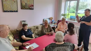Grupė įvairaus amžiaus žmonių sėdi prie stalo su raudona staltiese ir klausosi kalbėtojo.