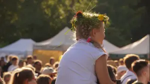 Maža mergaitė su gėlių vainiku ant galvos sėdi tėčio pečiais lauko renginyje.