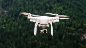 Baltas dronas skrenda virš miško, simbolizuojantis naująjį dronų kompetencijų centrą Druckūnuose.