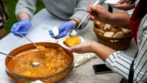 Moteris su pirštinėmis tepa džemą ant duonos riekelės prie stalo su maisto produktais.