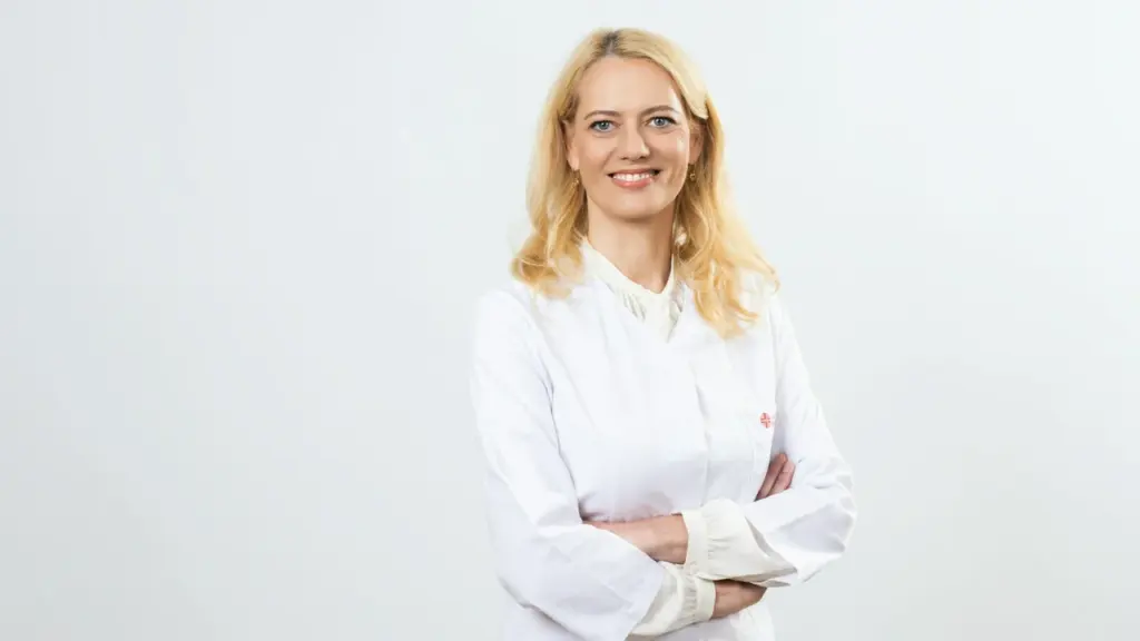 Šviesiaplaukė moteris gydytoja baltais chalatu, stovinti sukryžiuotomis rankomis šviesiame fone, žiūri į kamerą.