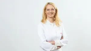 Šviesiaplaukė moteris gydytoja baltais chalatu, stovinti sukryžiuotomis rankomis šviesiame fone, žiūri į kamerą.