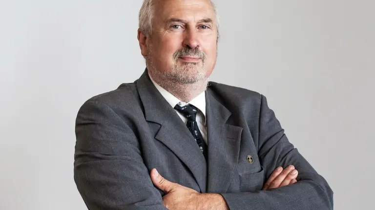 Vyresnio amžiaus vyras su akiniais ir kostiumu, stovintis sukryžiuotomis rankomis neutraliame fone.