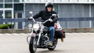 Vidutinio amžiaus vyras su motociklininko šalmu važiuoja juodu kreiserio tipo motociklu asfaltuota danga.
