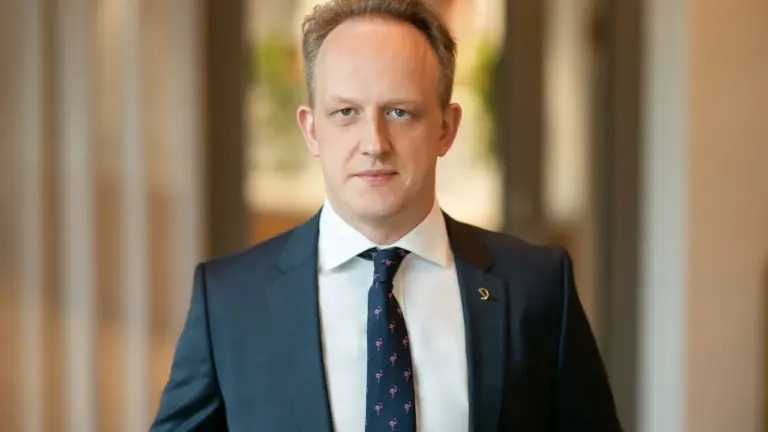 Vidutinio amžiaus vyras su tamsiu kostiumu ir margu kaklaraiščiu žvelgia į fotoobjektyvą.