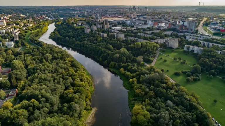 Neries upė vingiuoja per žalią mišką šalia daugiabučių namų ir Vilniaus miesto panoramos.