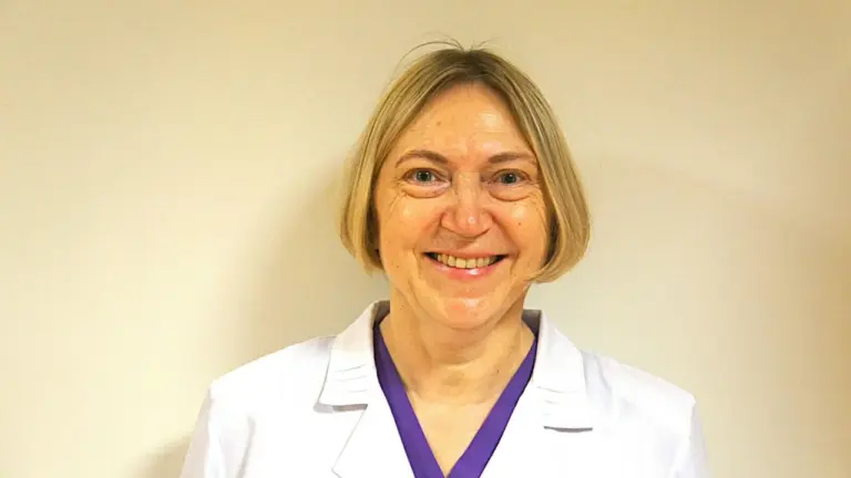 Besišypsanti vidutinio amžiaus moteris šviesiais plaukais, vilkinti baltą medicinos chalatą su violetine palaidine.