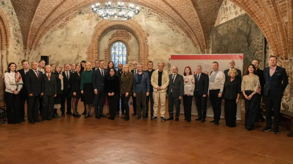 Didelė grupė oficialiai apsirengusių žmonių stovi istorinėje Trakų pilies menėje su arkinėmis lubomis.
