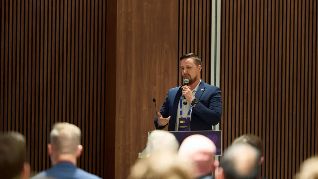 Barzdotas vyras su švarku kalba į mikrofoną prie tribūnos konferencijų salėje auditorijai.